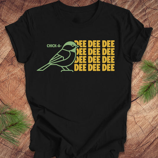 A black short-sleeve t-shirt laid flat on a rustic wood surface with pine branches in the corners. The shirt features a graphic of a green chickadee bird next to yellow text that reads 'CHICK-A-DEE DEE DEE DEE DEE DEE'.
