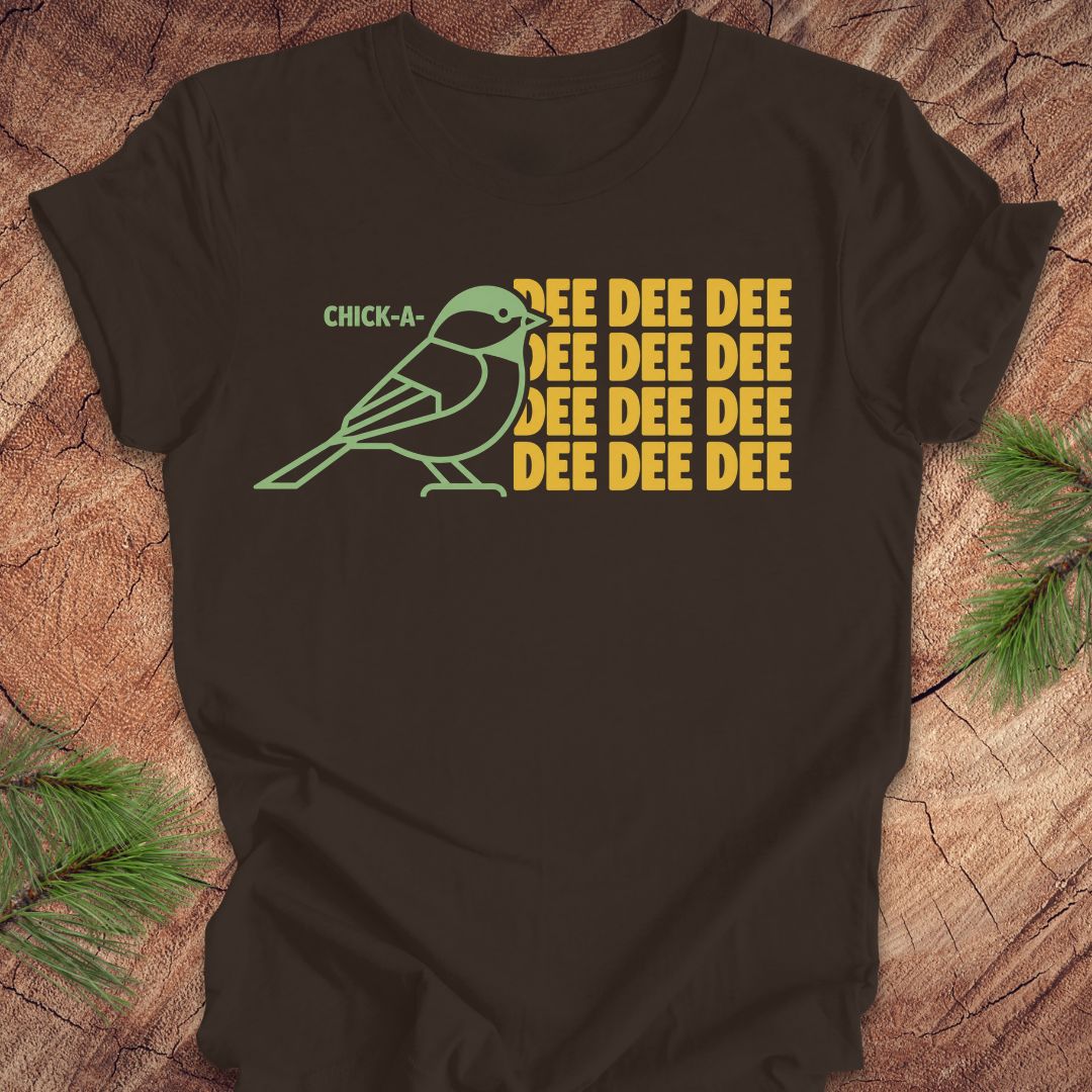 A brown short-sleeve t-shirt laid flat on a rustic wood surface with pine branches in the corners. The shirt features a graphic of a light green chick-a-doodle with the text 'CHICK-A' above it and 'DEE DEE DEE' repeated in yellow on the right.