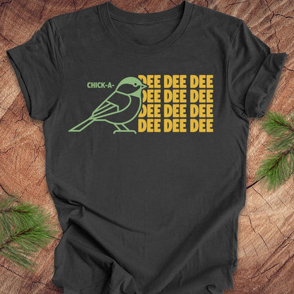 A dark gray short-sleeve t-shirt laid flat on a rustic wood surface with pine branches in the corners. The shirt features a graphic of a light green chick-a-doodle with the text 'CHICK-A' above it and 'DEE DEE DEE' repeated in yellow on the right.