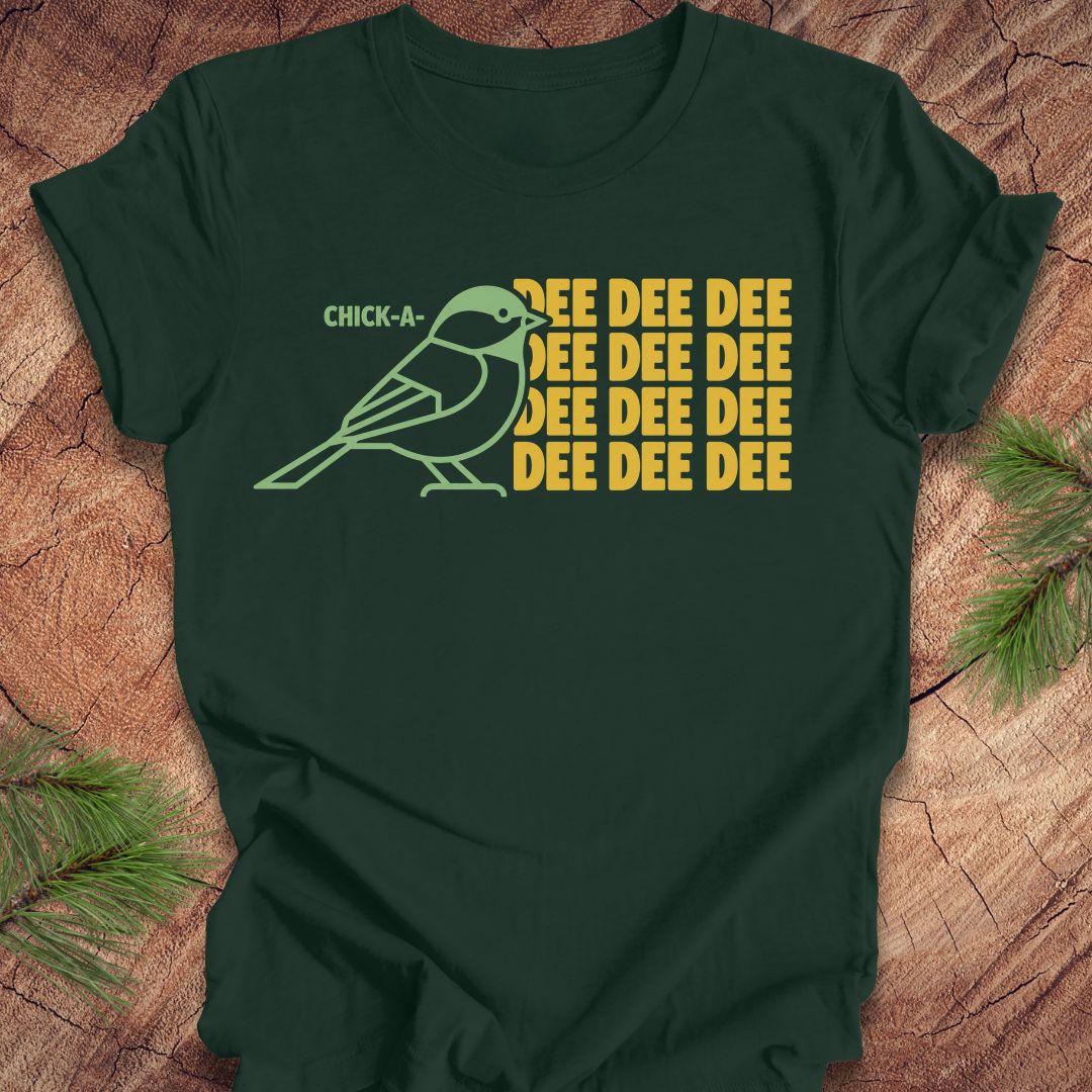 A dark forest green short-sleeve t-shirt laid flat on a rustic wood surface with pine branches in the corners. The shirt features a graphic of a light green chick-a-doodle with the text 'CHICK-A' above it and 'DEE DEE DEE' repeated in yellow on the right.