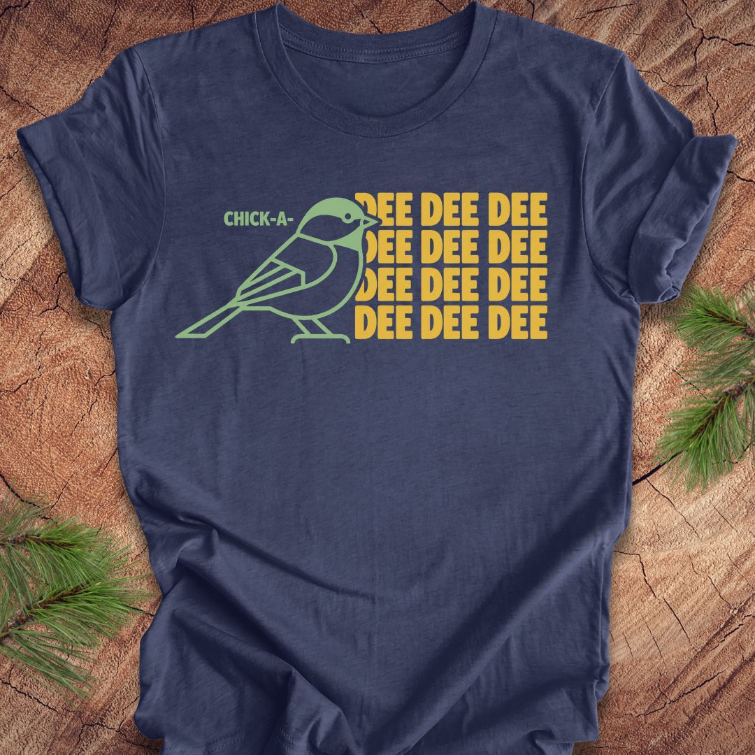 A navy blue short-sleeve t-shirt with a graphic print of a chick-a-mock bird and the text 'CHICK-A- MOCK' and 'DEE DEE DEE' in yellow, displayed flat on a rustic wood background with pine branches.