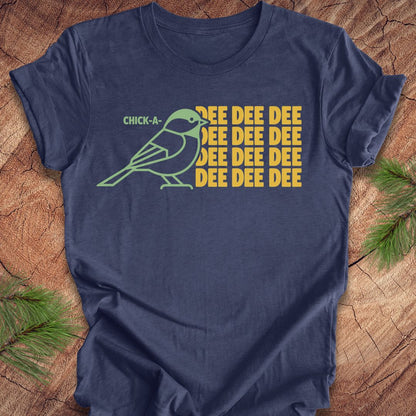 A navy blue short-sleeve t-shirt with a graphic print of a chick-a-mock bird and the text 'CHICK-A- MOCK' and 'DEE DEE DEE' in yellow, displayed flat on a rustic wood background with pine branches.