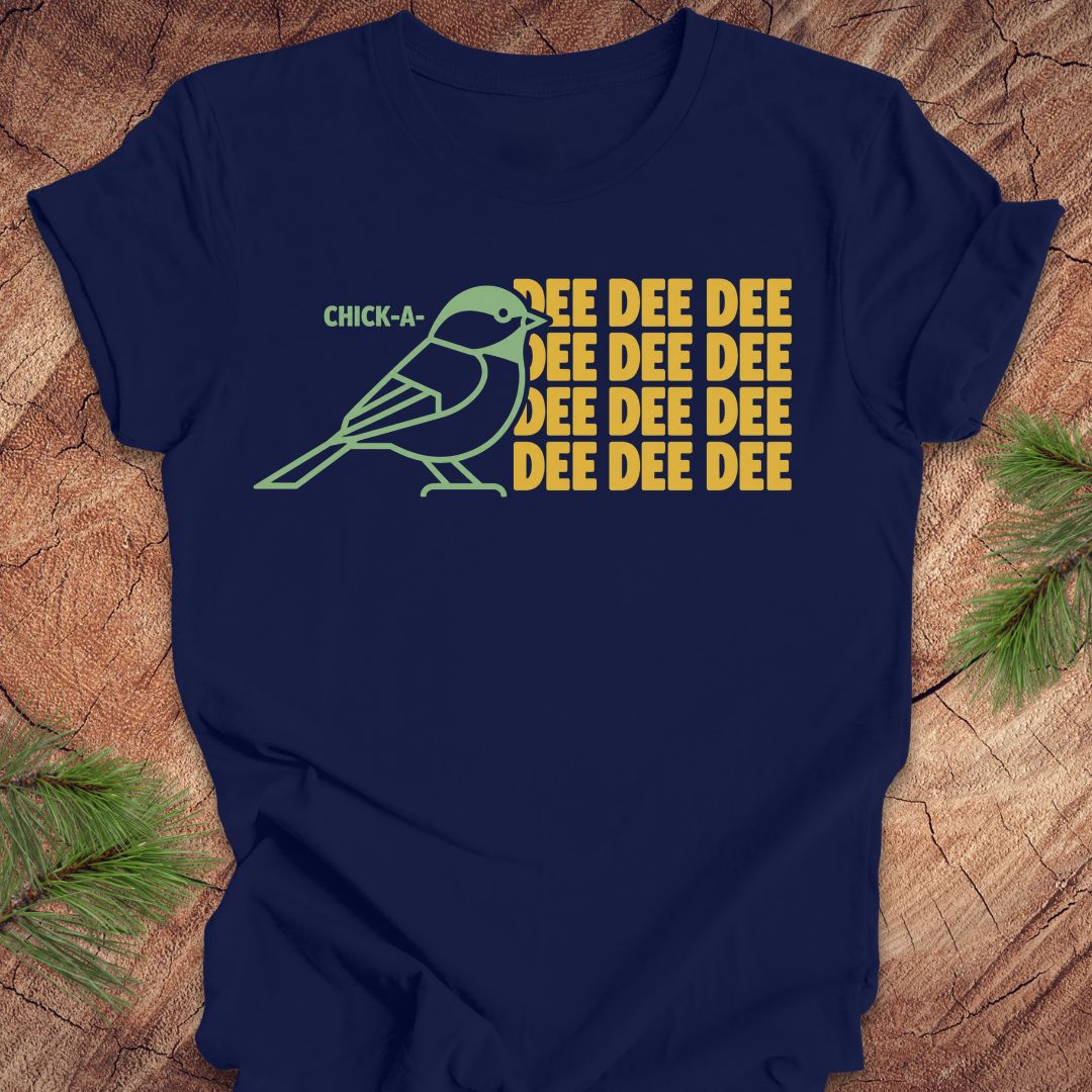 A navy blue short-sleeve t-shirt with a graphic print of a green chick-a-mock bird and the text 'CHICK-A' and 'DEE DEE DEE' in yellow, displayed flat on a rustic wood background with pine branches.