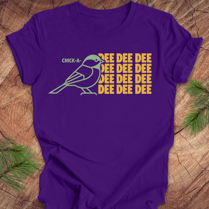 A purple short-sleeve crewneck t-shirt laid flat on a rustic wood background with pine branches in the corners. The shirt features a graphic of a light blue chick-a-doodle with the text 'CHICK-A' above it and 'DEE DEE DEE' repeated in yellow on the right.