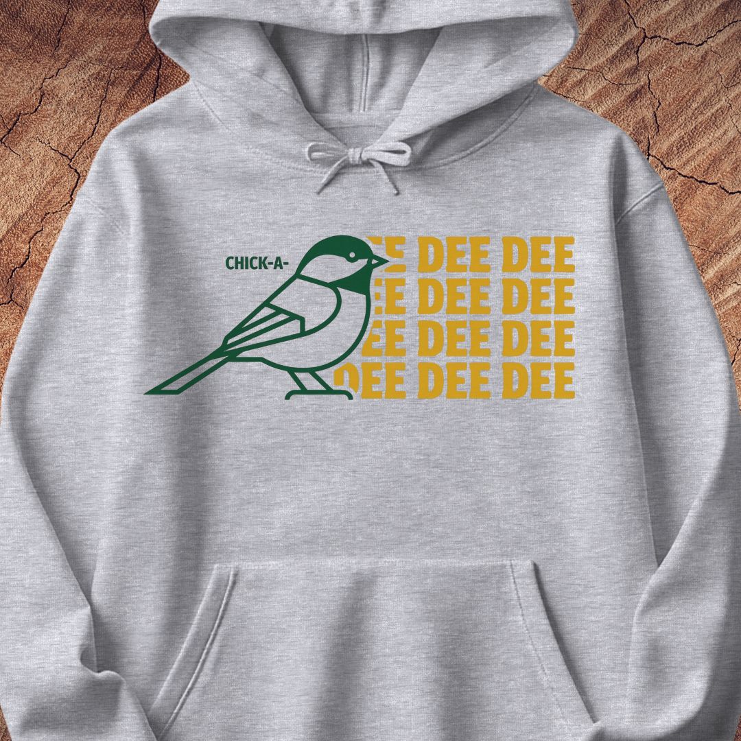 A light heather gray pullover hoodie featuring a graphic print of a green and white chickadee bird next to the text 'CHICK-A-DEE DEE DEE' in yellow, displayed flat against a rustic wood background.