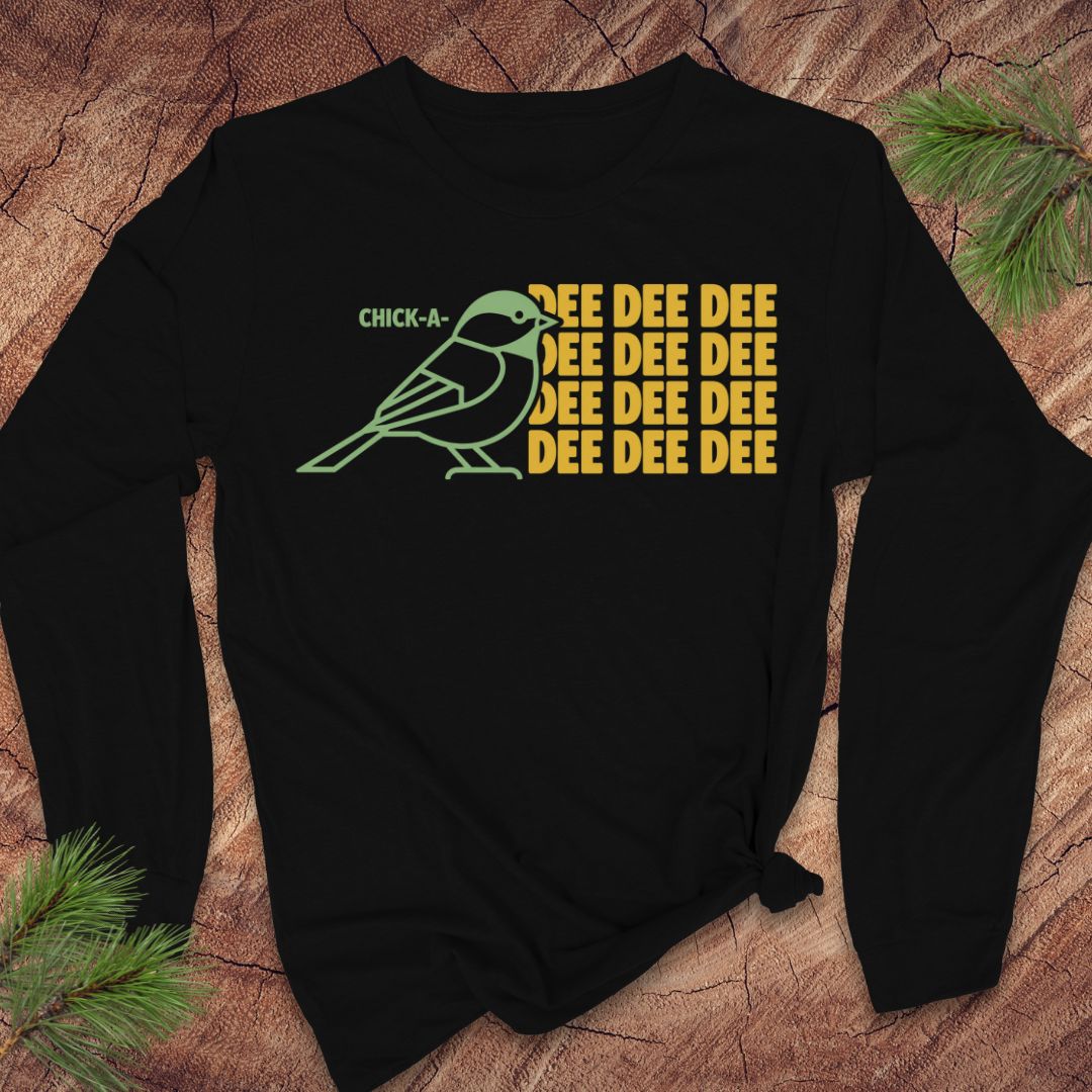 A black long-sleeve t-shirt with a graphic of a green chickadee and yellow text reading 'CHICK-A-DEE DEE DEE DEE DEE DEE', laid flat on a rustic wood background with pine branches.