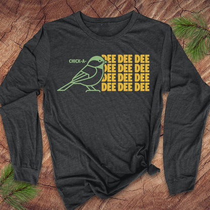 A dark charcoal gray long-sleeve t-shirt with a graphic print of a green chick-a-doodle and yellow text reading 'CHICK-A' and 'REE DEE DEE' repeated, laid flat on a rustic wood background with pine branches.
