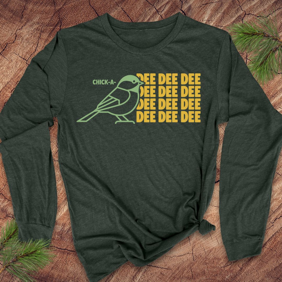 A dark forest green long-sleeve t-shirt with a graphic print of a chick-a-doodle and the text 'CHICK-A' and 'EEEEEEE', styled flat on a rustic wood background with pine needles.