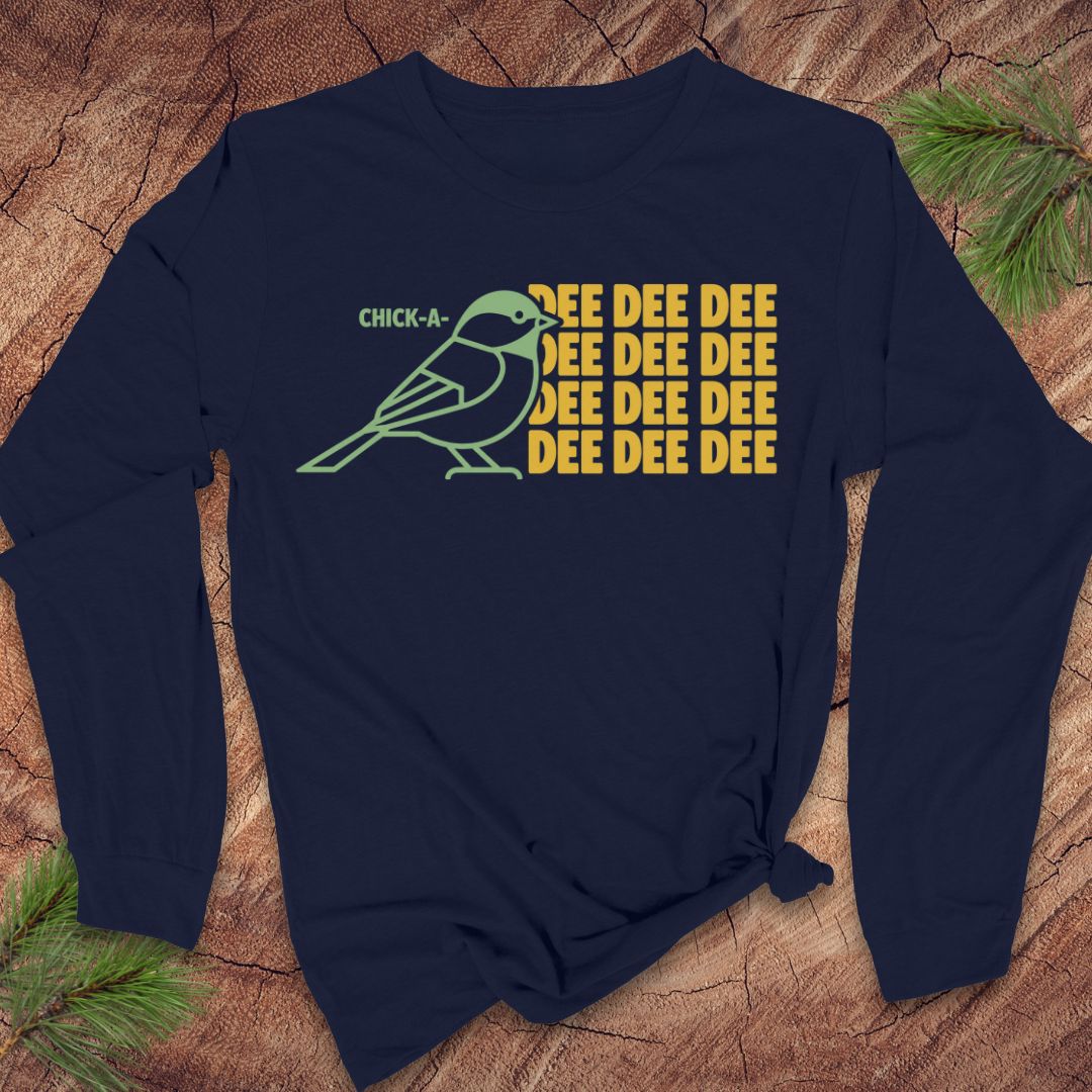 A navy long-sleeve t-shirt with a graphic of a chickadee and the text 'CHICK-A- DEE DEE DEE' in yellow and green, displayed flat on a rustic wood background with pine needles.