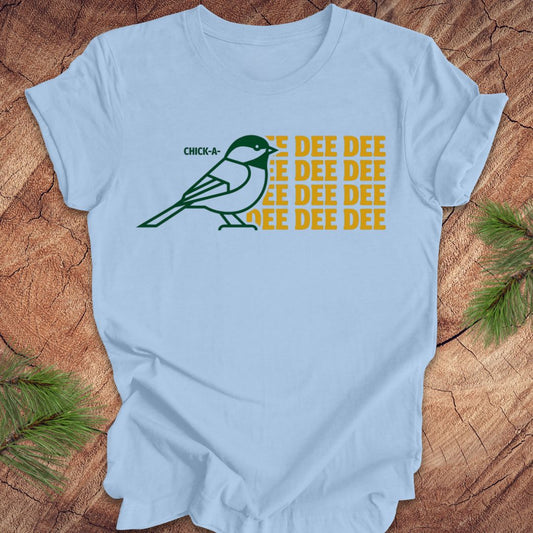 Light blue t-shirt with a green chickadee graphic and yellow text on a wooden surface.