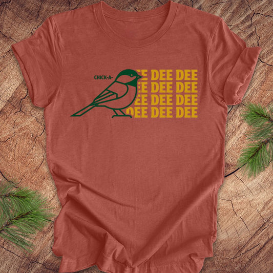 Clay colored t-shirt with a green chickadee graphic and yellow text on a wooden surface