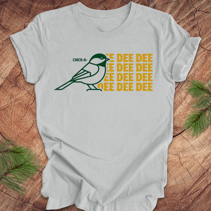 Silver t-shirt with a green chickadee graphic and yellow text on a wooden surface