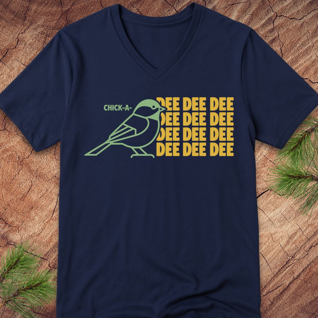 A navy blue V-neck t-shirt laid flat on a rustic wood background with pine branches in the corners. The shirt features a graphic of a green chickadee bird next to the text 'CHICK-A' and 'EEEEEEE' repeated in yellow.