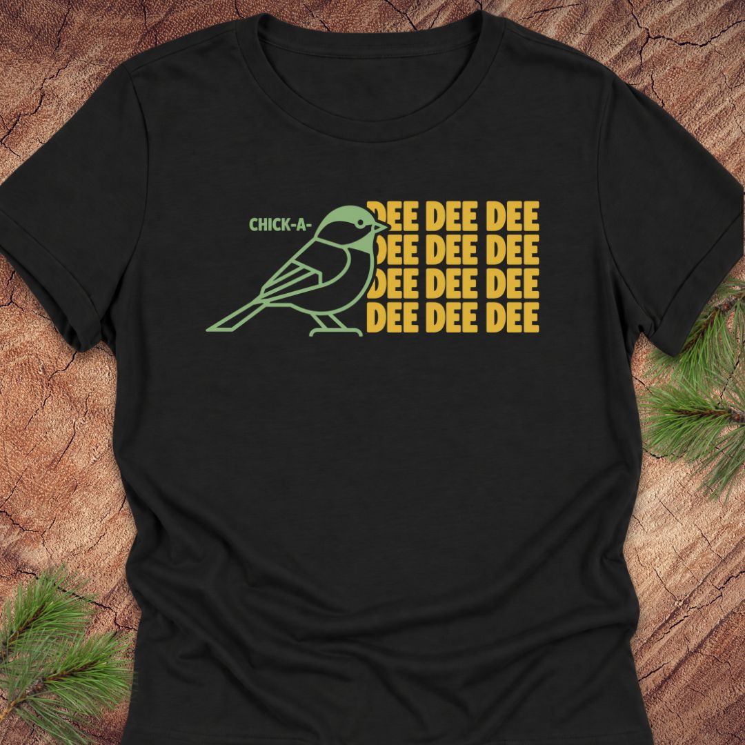 A black short-sleeve t-shirt with a graphic of a green chickadee and yellow text reading 'CHICK-A- SEE DEE DEE DEE DEE DEE DEE', laid flat on a rustic wood background with pine branches.
