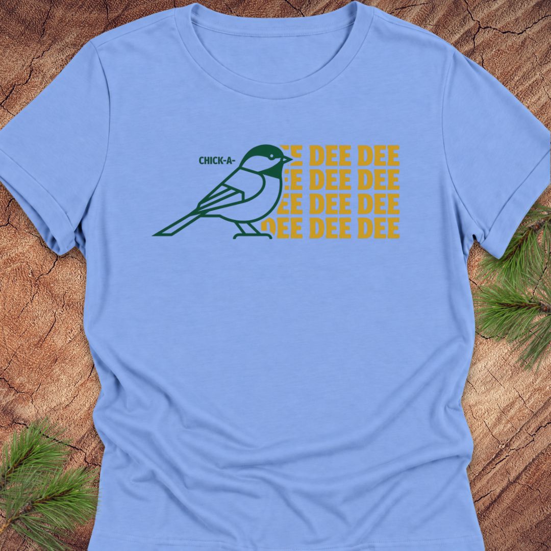 A light blue short-sleeve t-shirt laid flat on a rustic wood background with pine branches in the corners. The shirt features a graphic of a green chickadee bird next to yellow text that reads 'CHICK-A-DEE-DEE' repeated multiple times.