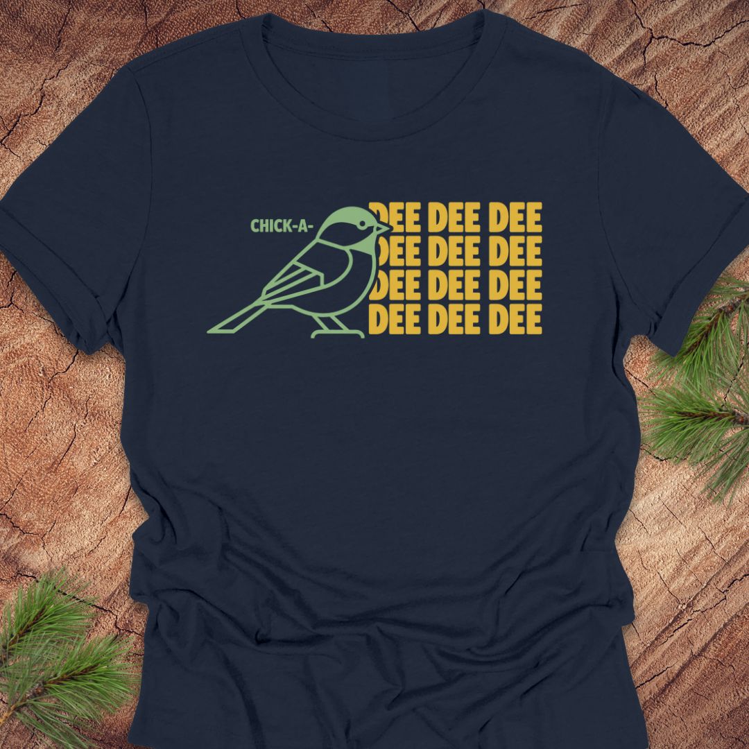 A navy blue short-sleeve t-shirt laid flat on a rustic wood background with pine branches in the corners. The shirt features a graphic of a green chick-a-doodle with the text 'CHICK-A-' above it and 'EEEE EEEE EEEE' repeated in yellow on the right.