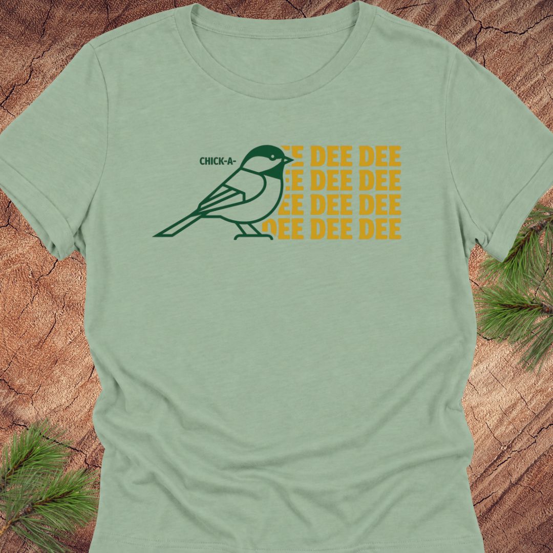 A sage green short-sleeve t-shirt with a graphic of a chickadee and the text 'CHICKADEE DEE DEE' in yellow and dark green, displayed flat on a rustic wood background with pine needles.