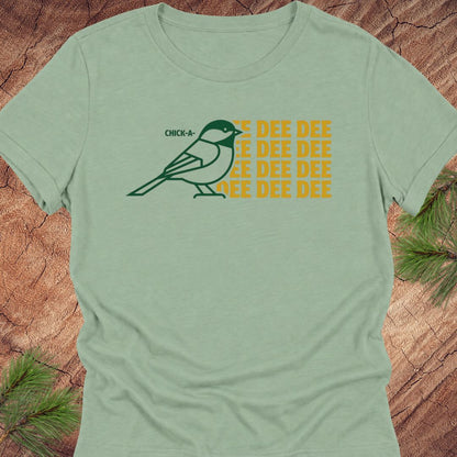 A sage green short-sleeve t-shirt with a graphic of a chickadee and the text 'CHICKADEE DEE DEE' in yellow and dark green, displayed flat on a rustic wood background with pine needles.
