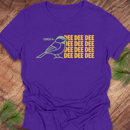 A purple short-sleeve t-shirt with a graphic of a blue and white bird next to yellow text reading 'CHICK-A-EEEEEEE', laid flat on a rustic wood background with pine branches.