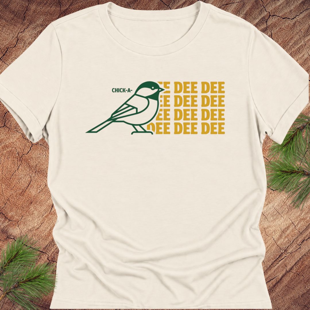 An off-white short-sleeve t-shirt with a graphic of a green chickadee and yellow text reading 'CHICK-A-DEE DEE DEE DEE DEE DEE', laid flat on a rustic wood background with pine branches.