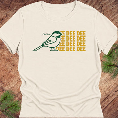 An off-white short-sleeve t-shirt with a graphic of a green chickadee and yellow text reading 'CHICK-A-DEE DEE DEE DEE DEE DEE', laid flat on a rustic wood background with pine branches.