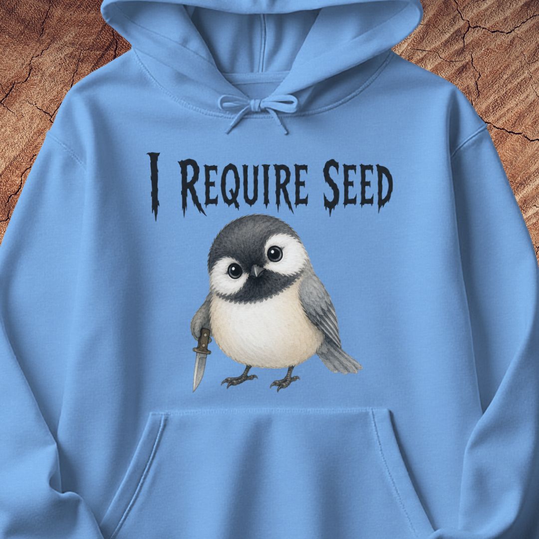 A light blue hoodie laid flat on a rustic wood background, featuring a printed graphic of a small bird holding a seed knife with the text 'I REQUIRE SEED' above it.