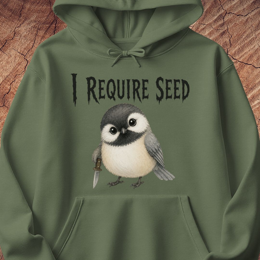 An olive green pullover hoodie featuring a graphic print of a small bird holding a seed knife, with the text 'I REQUIRE SEED' above it, displayed against a textured wood background.