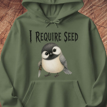 An olive green pullover hoodie featuring a graphic print of a small bird holding a seed knife, with the text 'I REQUIRE SEED' above it, displayed against a textured wood background.