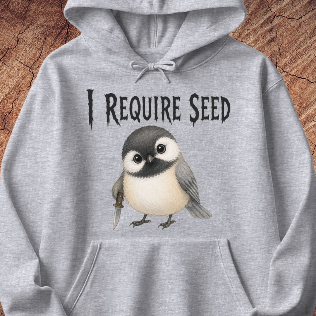 A light heather gray pullover hoodie featuring a graphic print of a small bird holding a dagger beneath the text 'I REQUIRE SEED' in a gothic font, displayed against a rustic wood background.