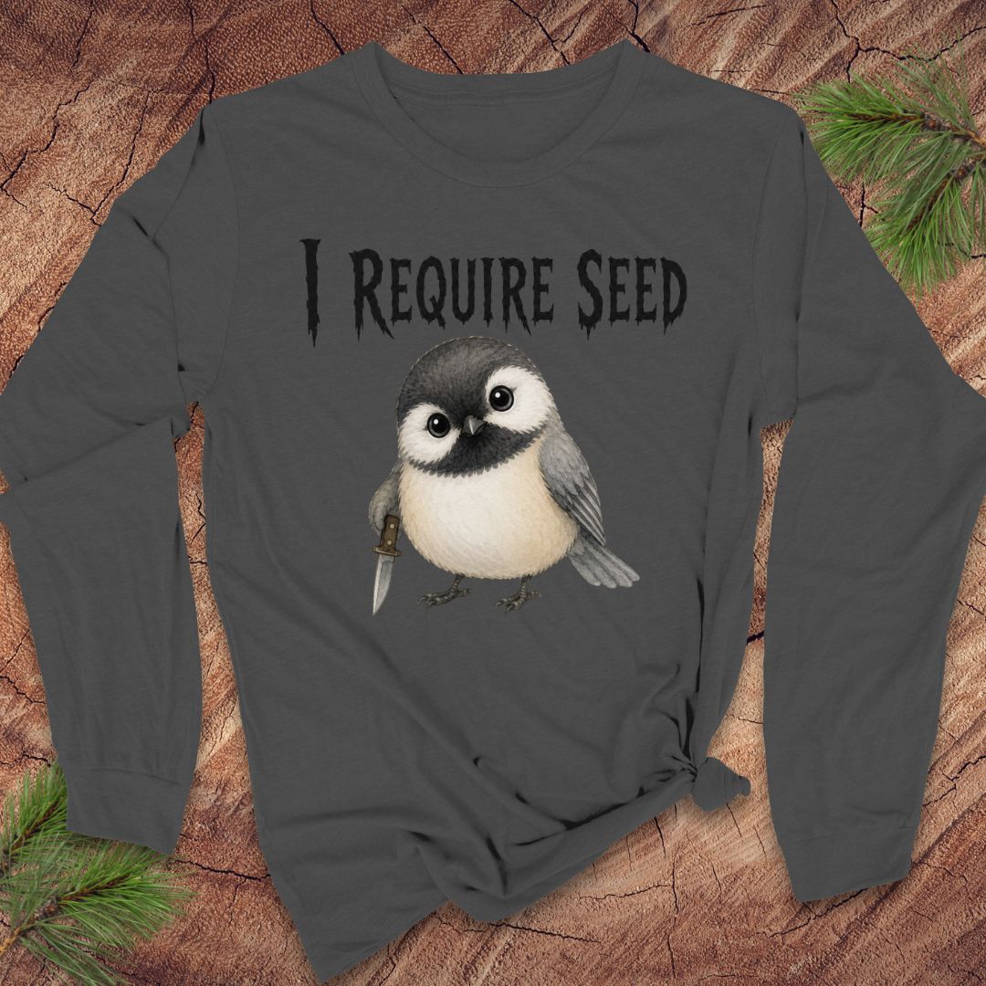 A dark gray long-sleeve t-shirt with a graphic of a small bird and the text 'I REQUIRE SEED' printed on the front, styled on a rustic wood background with pine needles.