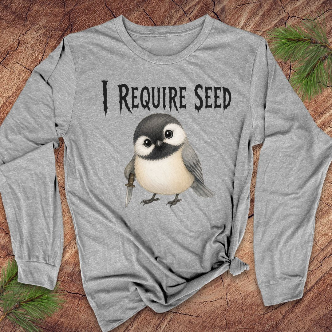 A heather gray long-sleeve t-shirt with a graphic of a cute bird and the text 'I REQUIRE SEED' printed on the chest, laid flat on a rustic wood background with pine needles.