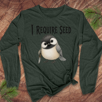 A dark forest green long-sleeve t-shirt with a graphic of a chickadee bird and the text 'I REQUIRE SEED' printed on the front, styled flat on a rustic wood background with pine needles.