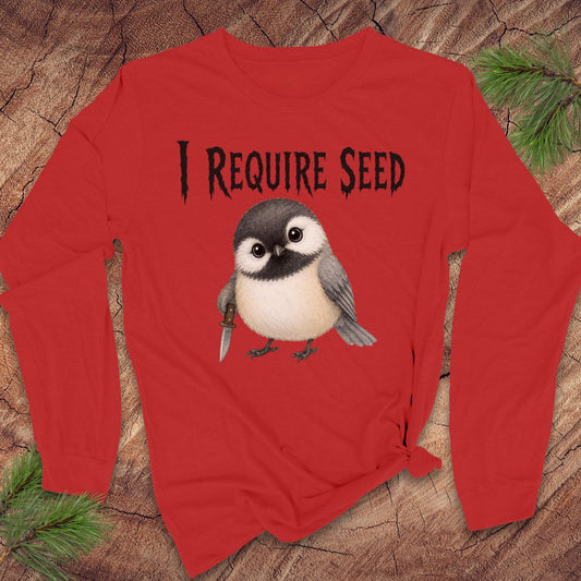 A red long-sleeve t-shirt with a graphic of a bird holding a seed pod and the text 'I REQUIRE SEED' in black, displayed flat on a rustic wood background with pine branches.