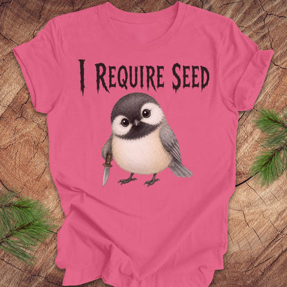 An bright pink short-sleeve T-shirt laid flat on a wood surface with autumn leaves around it. The shirt has black text reading 'I REQUIRE SEED' above a cartoon chickadee holding a dagger.