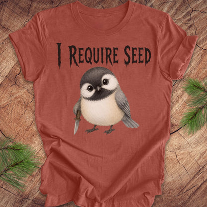 A terracotta colored short-sleeve T-shirt laid flat on a wood surface with autumn leaves around it. The shirt has black text reading 'I REQUIRE SEED' above a cartoon chickadee holding a dagger.