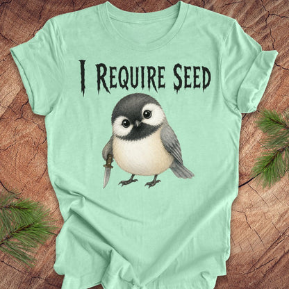 An mint green short-sleeve T-shirt laid flat on a wood surface with autumn leaves around it. The shirt has black text reading 'I REQUIRE SEED' above a cartoon chickadee holding a dagger.