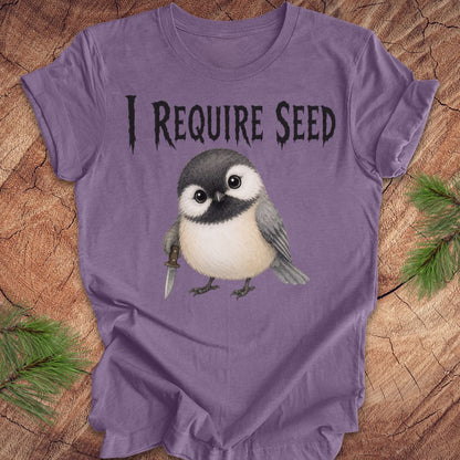 An purple short-sleeve T-shirt laid flat on a wood surface with autumn leaves around it. The shirt has black text reading 'I REQUIRE SEED' above a cartoon chickadee holding a dagger.