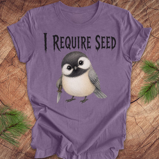 An purple short-sleeve T-shirt laid flat on a wood surface with autumn leaves around it. The shirt has black text reading 'I REQUIRE SEED' above a cartoon chickadee holding a dagger.