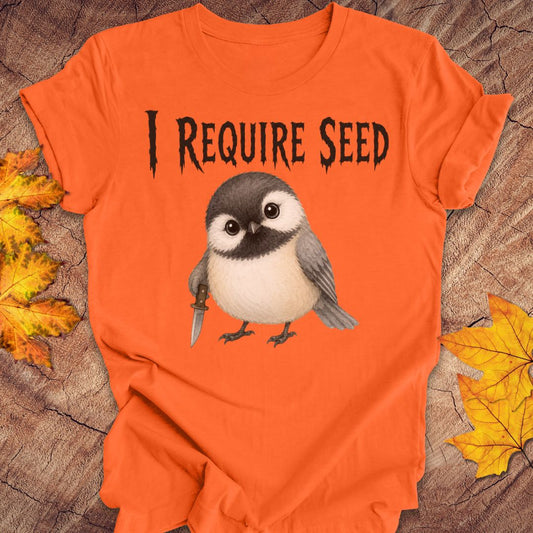 An orange short-sleeve T-shirt laid flat on a wood surface with autumn leaves around it. The shirt has black text reading 'I REQUIRE SEED' above a cartoon chickadee holding a dagger.