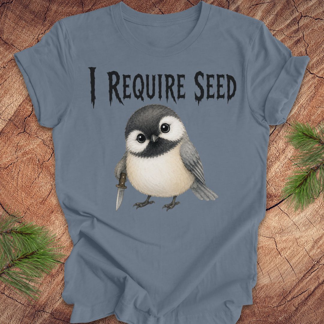 A slate blue short-sleeve T-shirt laid flat on a wood surface with autumn leaves around it. The shirt has black text reading 'I REQUIRE SEED' above a cartoon chickadee holding a dagger.