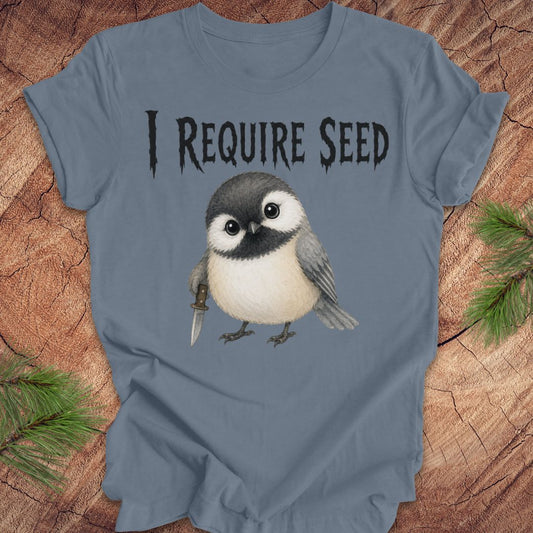 A slate blue short-sleeve T-shirt laid flat on a wood surface with autumn leaves around it. The shirt has black text reading 'I REQUIRE SEED' above a cartoon chickadee holding a dagger.