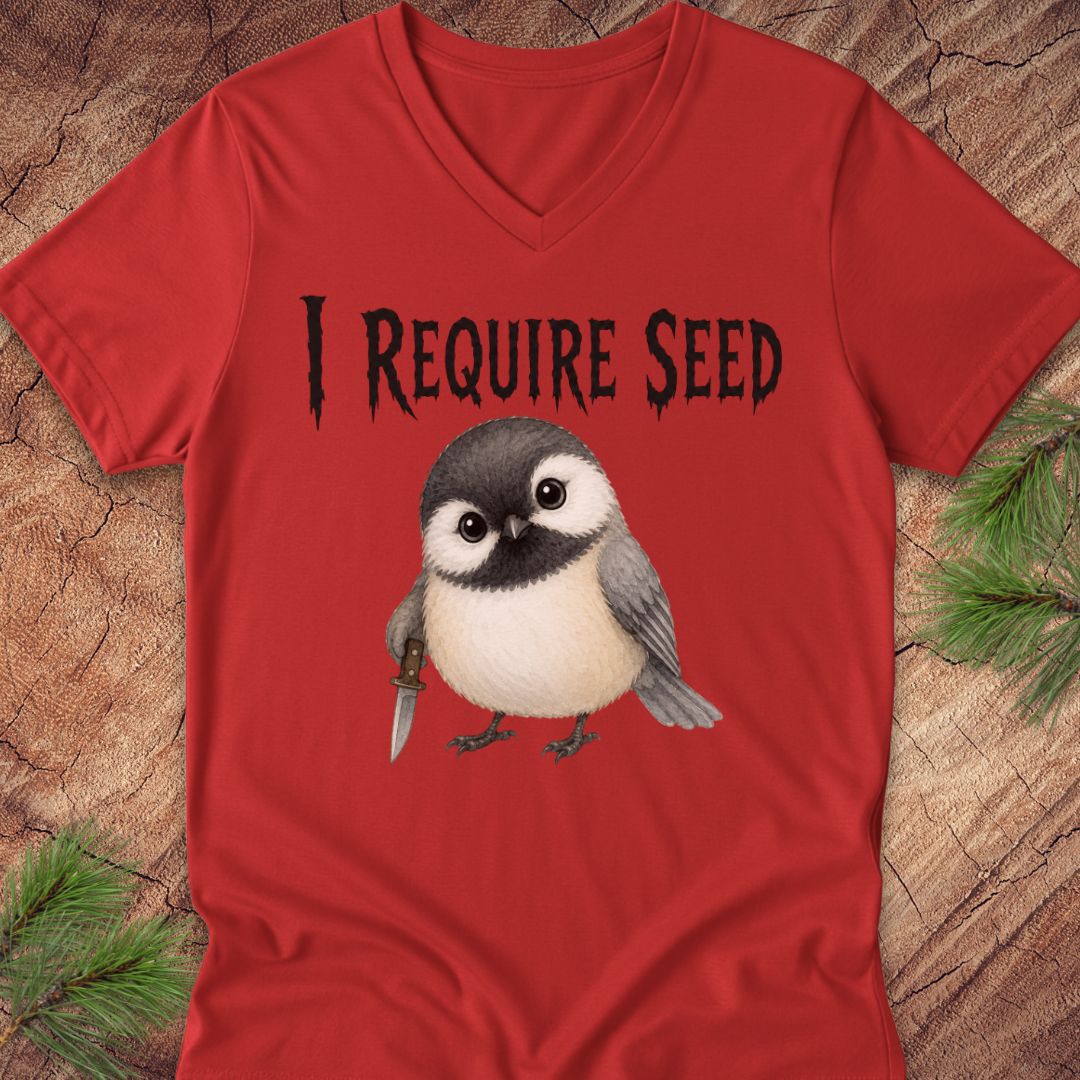 A red V-neck t-shirt laid flat on a rustic wood background with pine branches in the corners. The shirt features a graphic of a chickadee holding a knife and the text 'I REQUIRE SEED' in a black gothic font.