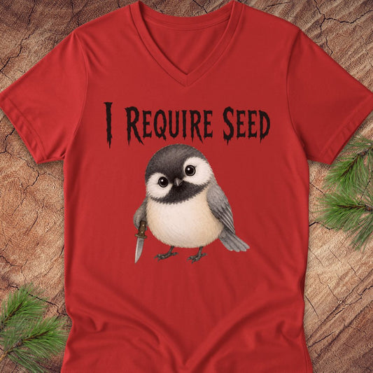 A red V-neck t-shirt laid flat on a rustic wood background with pine branches in the corners. The shirt features a graphic of a chickadee holding a knife and the text 'I REQUIRE SEED' in a black gothic font.