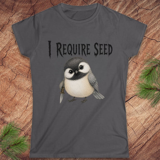 A dark gray short-sleeve t-shirt with a graphic of a cute chickadee holding a dagger and the text 'I REQUIRE SEED' in a gothic font, displayed flat on a rustic wood background with pine needles.