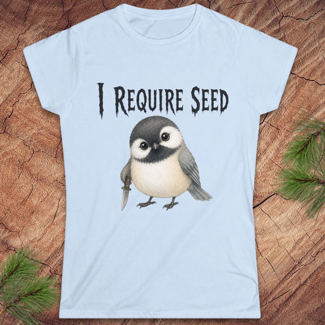 A light blue short-sleeve t-shirt with a graphic of a cute chickadee holding a dagger and the text 'I REQUIRE SEED' in a gothic font, displayed flat on a rustic wood background with pine needles.