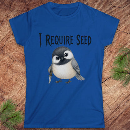 A royal blue short-sleeve t-shirt with a graphic of a cute chickadee holding a dagger and the text 'I REQUIRE SEED' in a gothic font, displayed flat on a rustic wood background with pine needles.