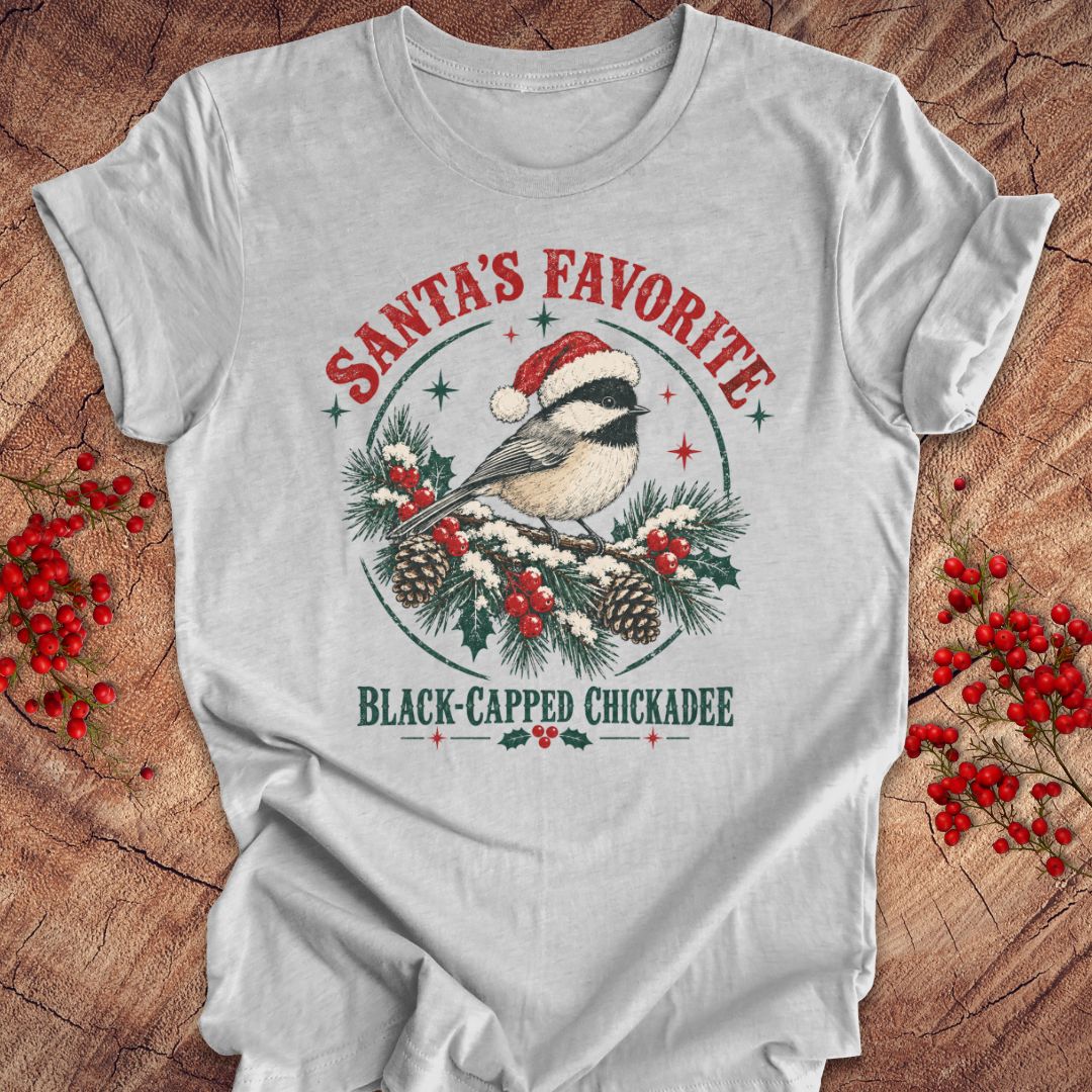 Athletic heather gray t-shirt with a Black-capped Chickadee in a Santa hat on a snowy pine bough with holly and red berries, 'Santa's Favorite' and 'Black-capped Chickadee' text, laid flat on a rustic wood background with red winter berries.