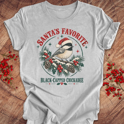Athletic heather gray t-shirt with a Black-capped Chickadee in a Santa hat on a snowy pine bough with holly and red berries, 'Santa's Favorite' and 'Black-capped Chickadee' text, laid flat on a rustic wood background with red winter berries.
