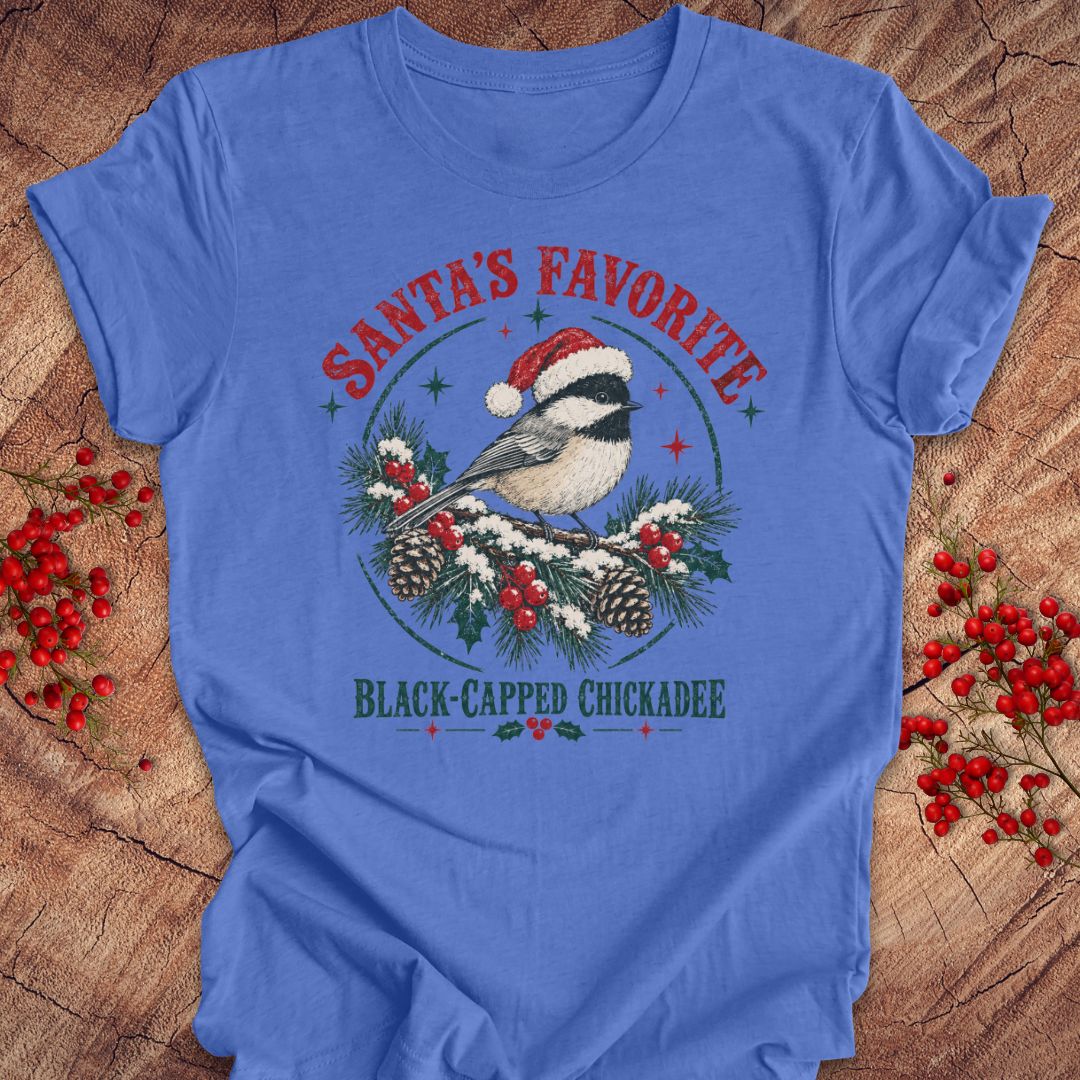 Heather Columbia blue t-shirt with a Black-capped Chickadee in a Santa hat on a snowy pine bough with holly and red berries, 'Santa's Favorite' and 'Black-capped Chickadee' text, laid flat on a rustic wood background with red winter berries.
