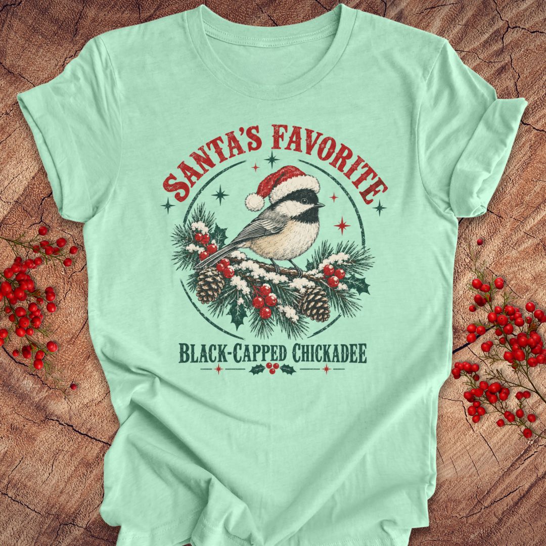 Heather mint green t-shirt with a Black-capped Chickadee in a Santa hat on a snowy pine bough with holly and red berries, 'Santa's Favorite' and 'Black-capped Chickadee' text, laid flat on a rustic wood background with red winter berries.